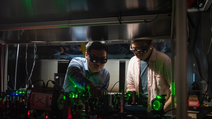 Two men wear safety glasses and look at an optical apparatus consisting of many lenses and mirrors and prisms in the foreground.