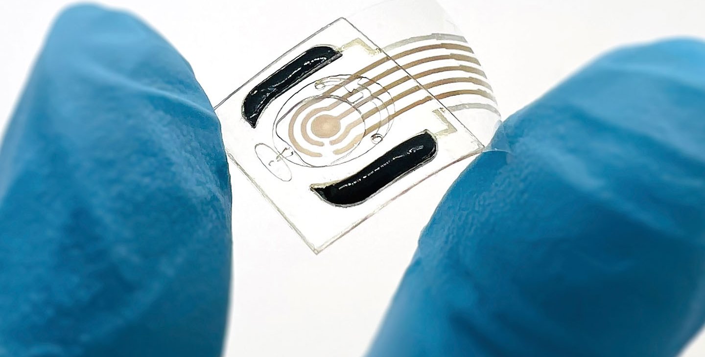 A hand wearing a blue rubber glove holds a semi-transparent plastic film containing a sensor between its index finger and thumb.
