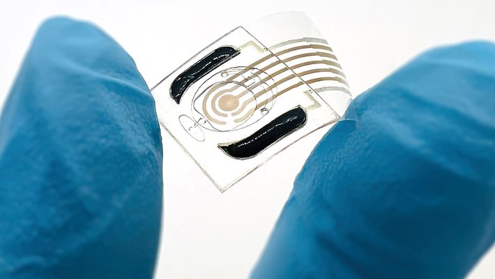 A hand wearing a blue rubber glove holds a semi-transparent plastic film containing a sensor between its index finger and thumb.