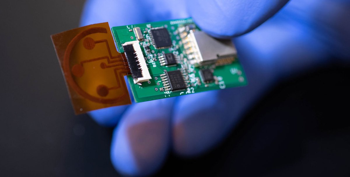 A hand wearing a blue rubber glove holds a small sensor that consists of a circuit board and a thin piece of plastic film.