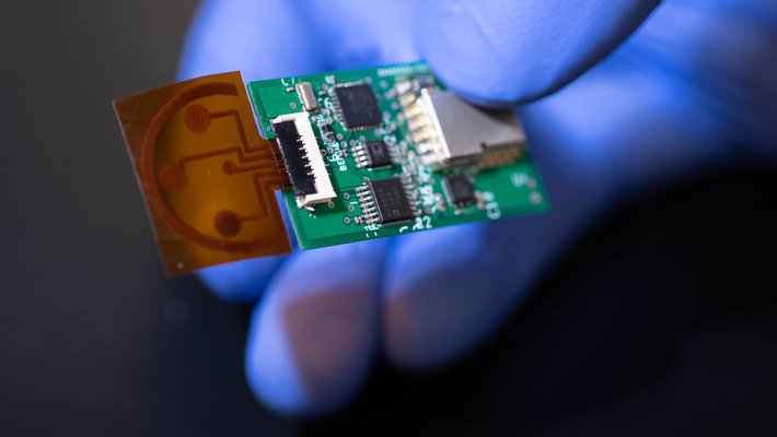 A hand wearing a blue rubber glove holds a small sensor that consists of a circuit board and a thin piece of plastic film.
