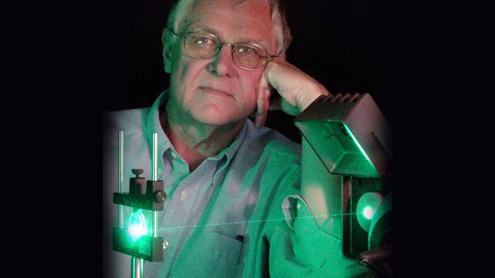 Portrait of an older man with gray hair and glasses posed behind a laser instrument shining green light