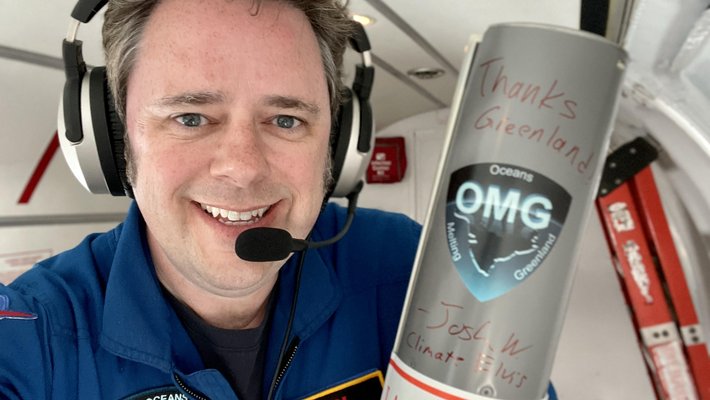 Photo of JPL researcher Josh Willis aboard a small aircraft wearing a flightsuit and holding a cylindrical launching tube used in the OMG mission.