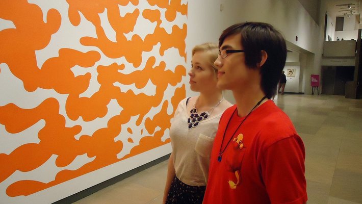 A couple looks at a piece of art in the Phoenix Art Museum. They are a young man and young woman who look at a modernist painting resembling orange clouds.