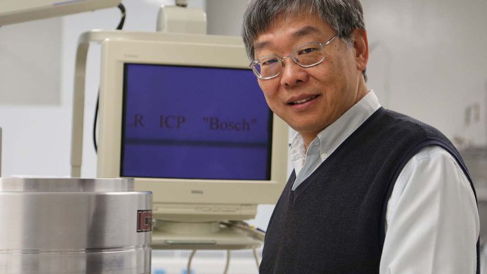 Yu-Chong "Y.C." Tai stands in a laboratory smiling at the camera. A computer monitor is in the background.
