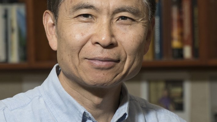 A portrait of Zhen-Gang Wang. He wears a dress shirt and appears in front of shelves of books.