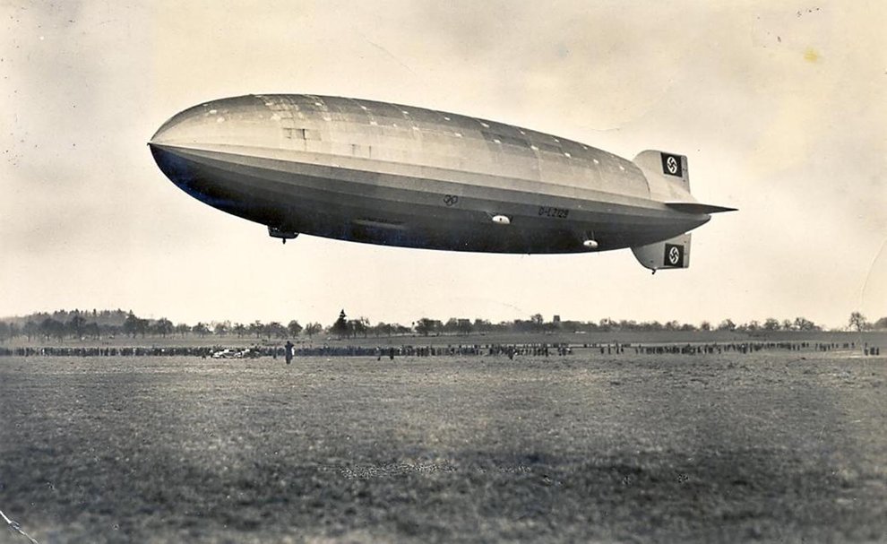 A sepia-toned black-and-white photograph of the Hindenburg hovering over a field. Spectators gathered below are dwarfed by its size.