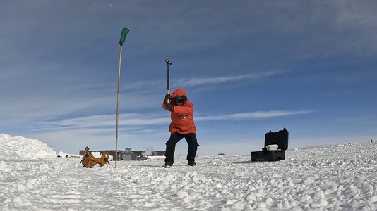 Zhongwen Zhan in a bright red winter coat swinging a sledgehammer at the Antarctic ice