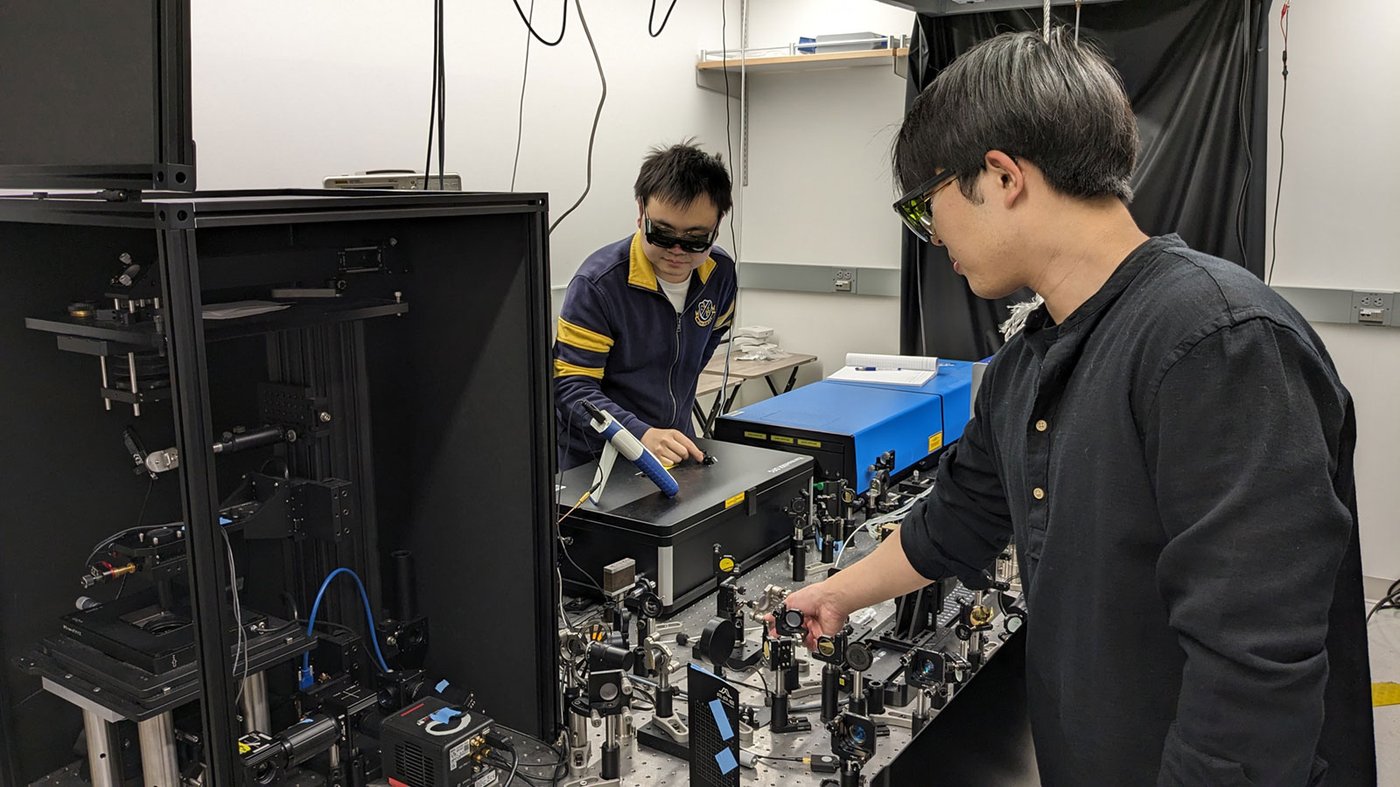 Postodoctoral scholar Haomin Wang (left) and graduate student Dongkwan Lee (right) demonstrate operation of the BonFIRE microscopy apparatus. It consists of a collection of optical devices like lenses attached to a table.