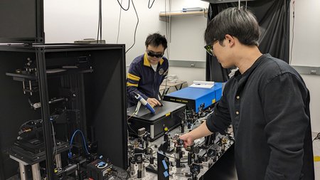 Postodoctoral scholar Haomin Wang (left) and graduate student Dongkwan Lee (right) demonstrate operation of the BonFIRE microscopy apparatus. It consists of a collection of optical devices like lenses attached to a table.
