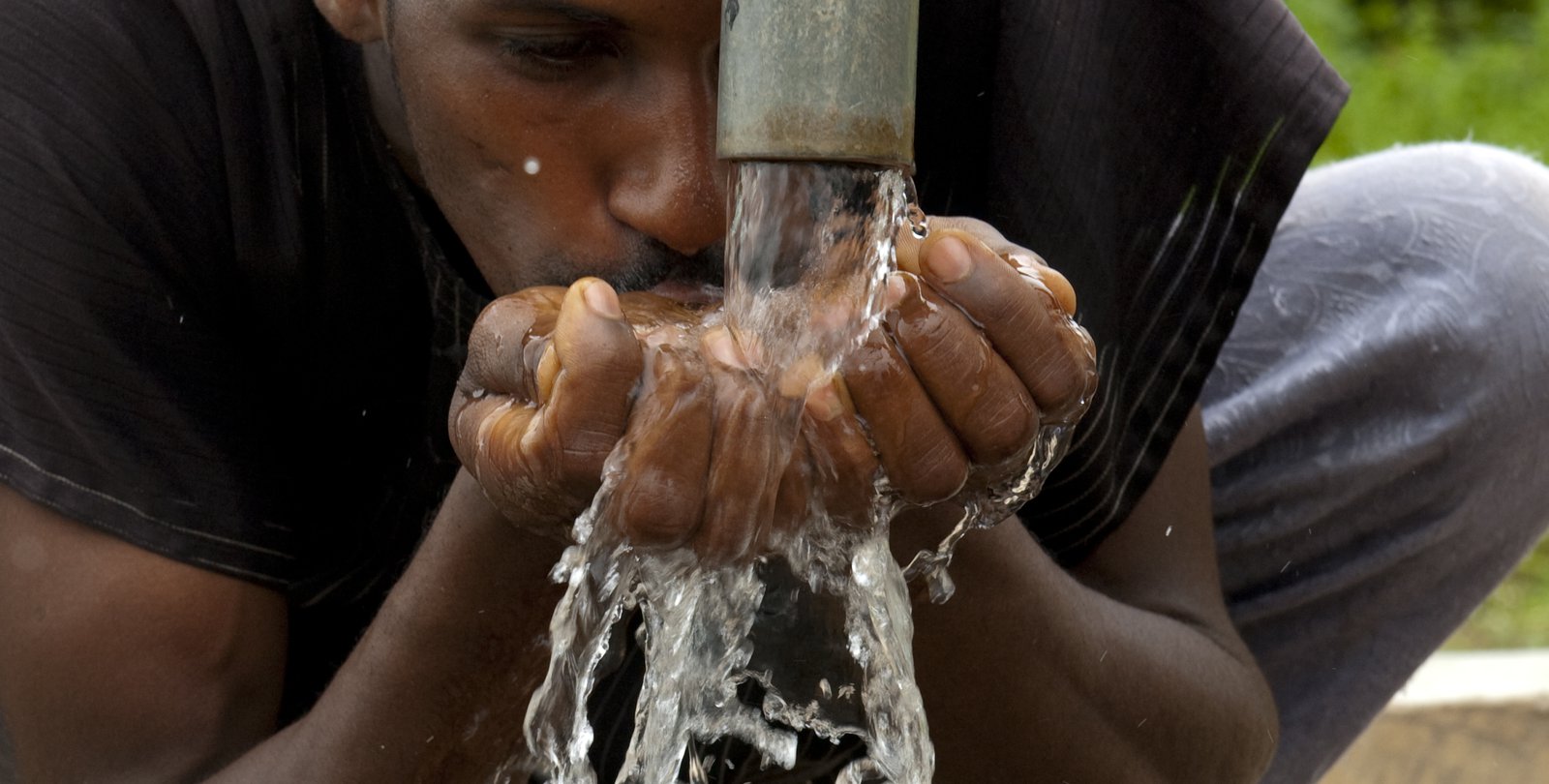 A man cups his hands under a pipe to catch and drink water.