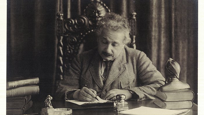 Einstein writes by hand at his desk. A bust of Dante sits on the corner of the desk near some books.