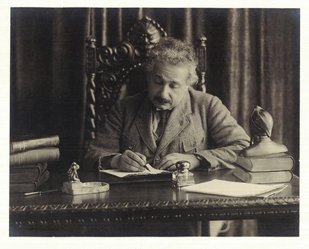 Einstein writes by hand at his desk. A bust of Dante sits on the corner of the desk near some books.