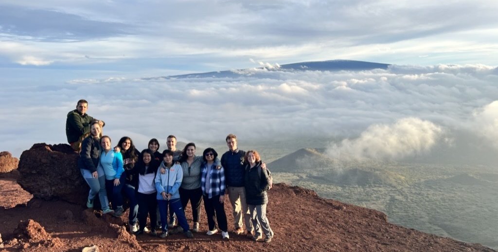 12 people stand together on soil and rocks with a green plain and clouds behind them