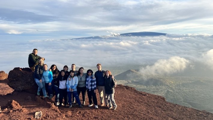 12 people stand together on soil and rocks with a green plain and clouds behind them