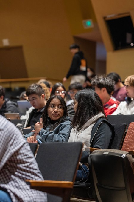 Two students sitting in a lecture hall talk to one another
