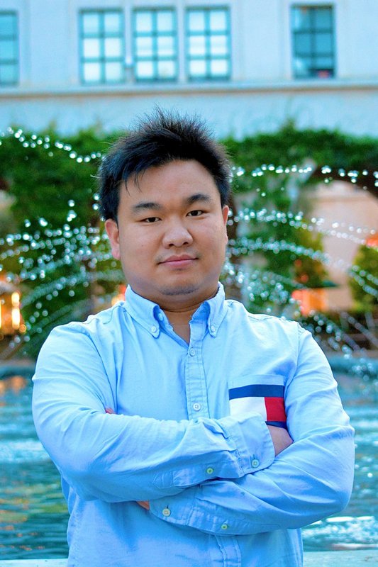 A portrait of Rui Cao, who stands in front of a fountain with his arms crossed.